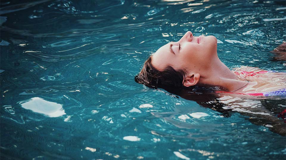 Auf dem Bild ist eine Frau im Schwimmbad zu sehen, umgeben von glitzerndem, klarem Wasser. Sie trägt einen bunten Badeanzug, der ihre Figur betont und ihre lebendige Persönlichkeit widerspiegelt.